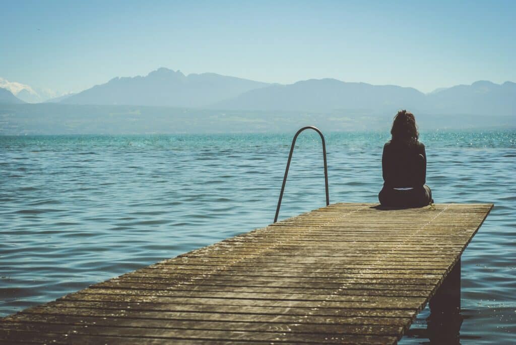 Person sitting alone on a dock overlooking a calm lake and mountains.